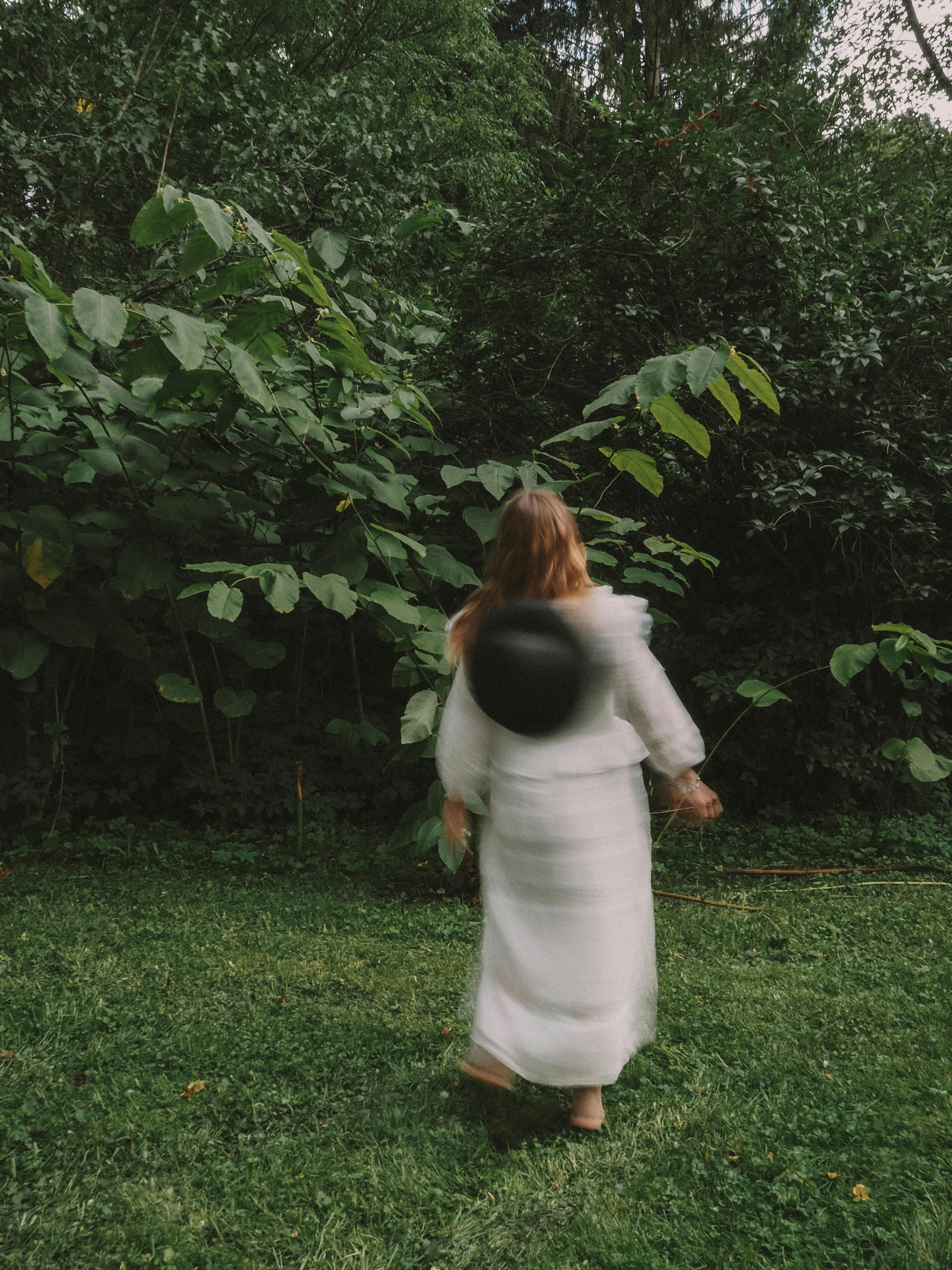 A blurry woman in a white dress walks through lush greenery, creating a carefree mood.