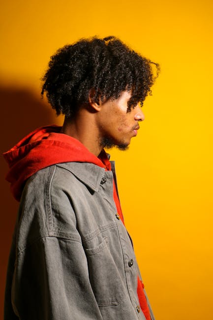 Side profile of a young man wearing a red hoodie and denim jacket against a vibrant yellow background.