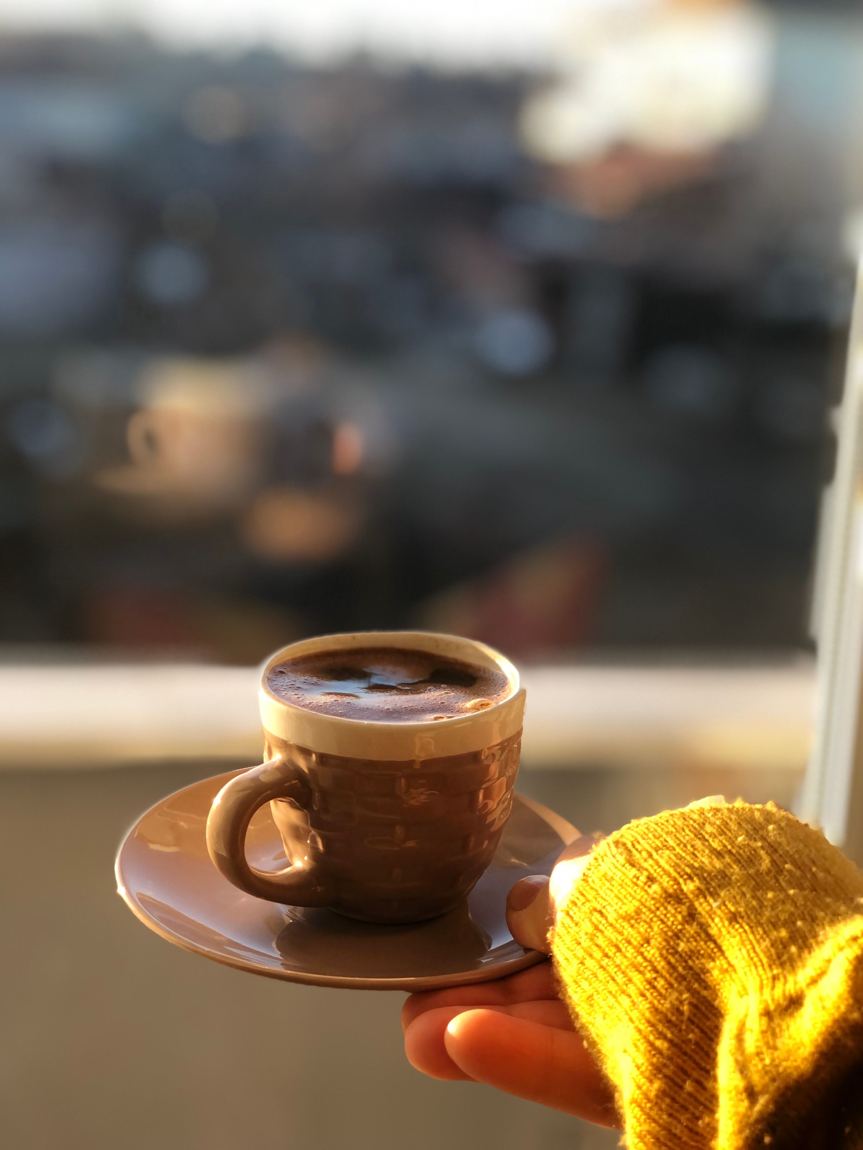 Free A person holding a steaming cup of coffee with a blurred city view in the background. Stock Photo