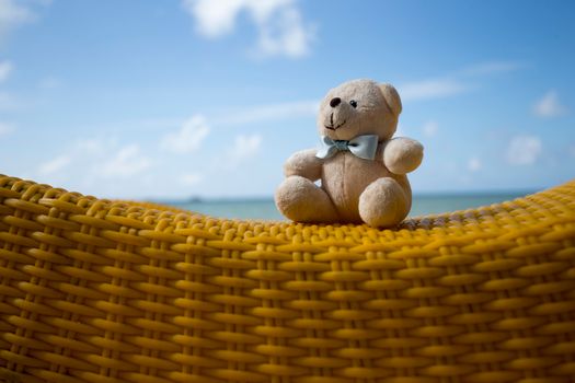 Adorable teddy bear with bowtie sitting on yellow woven seat under blue sky.