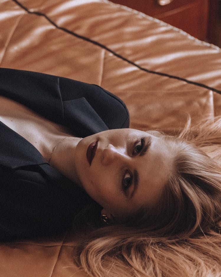 Blond Woman With Long Hair Laying In Black Jacket On Bed 