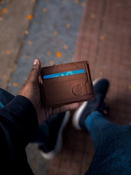 A close-up view of a man's hand holding a brown wallet with a credit card visible, captured on a sidewalk.