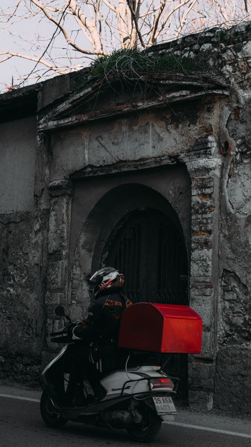 Free Motorcyclist with helmet and delivery box rides past an old stone building in an urban setting. Stock Photo
