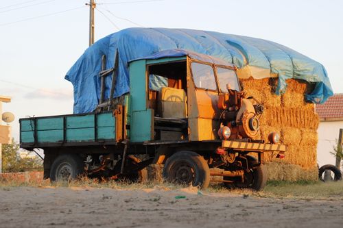Free Old truck parked outdoors next to hay bales under a blue tarp in rural countryside. Stock Photo