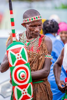 An African man in traditional attire holding a shield at a cultural event.