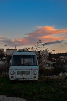 A vintage van parked on grass against a backdrop of city buildings under a colorful sunset sky.