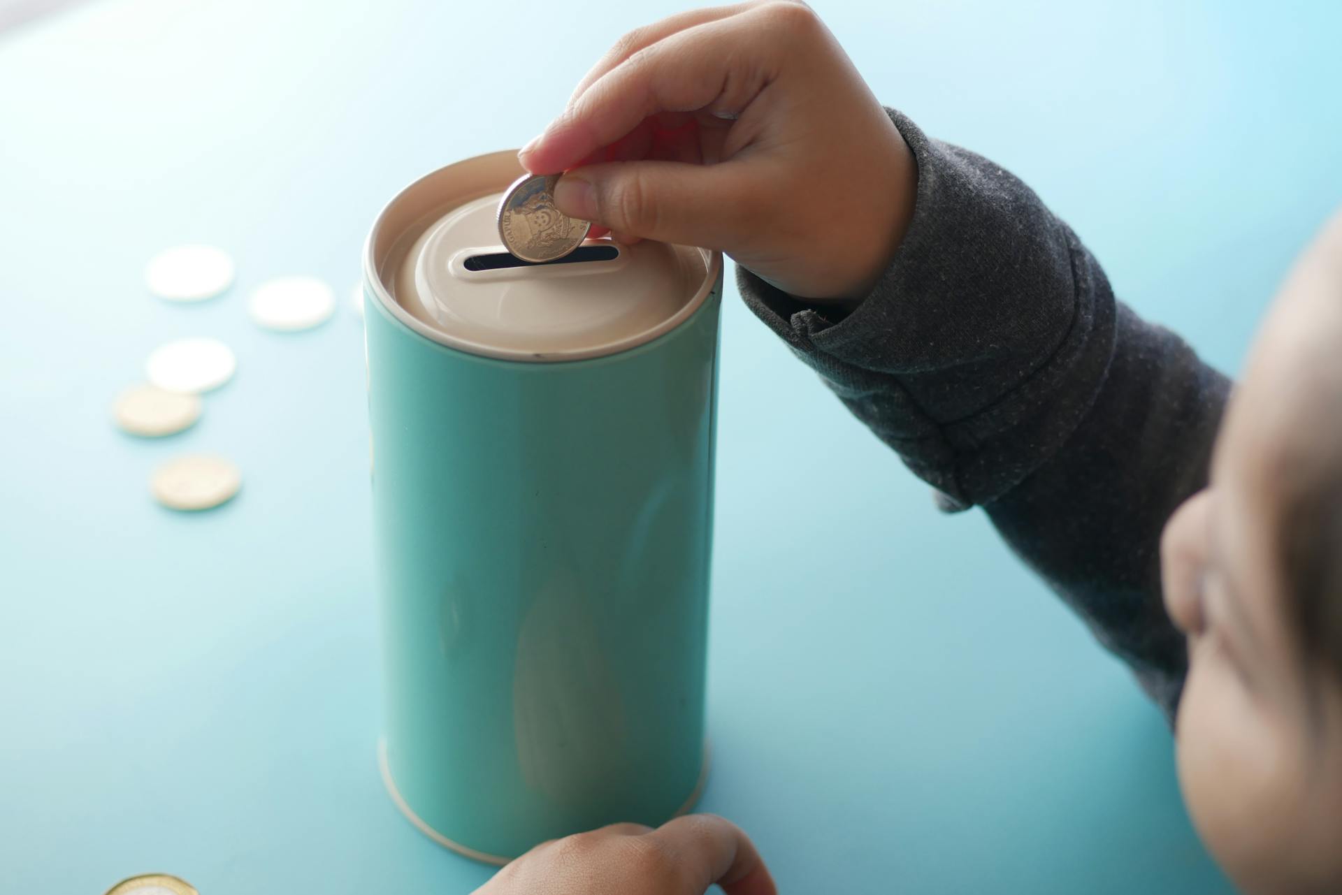 A child placing a coin into a teal piggy bank, representing saving money and financial education.