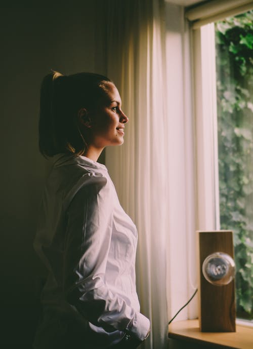 Free Woman Wearing White Long-sleeved Shirt Standing in Front of the Window With White Curtain Stock Photo