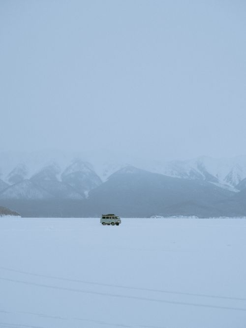Free A solitary van travels across a vast, snow-covered mountain landscape in winter. Stock Photo