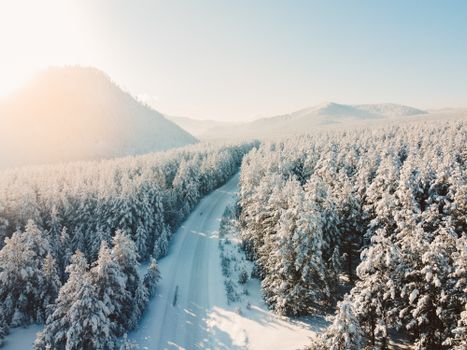 A stunning aerial shot of a snow-covered road cutting through a winter forest with mountains in the background.