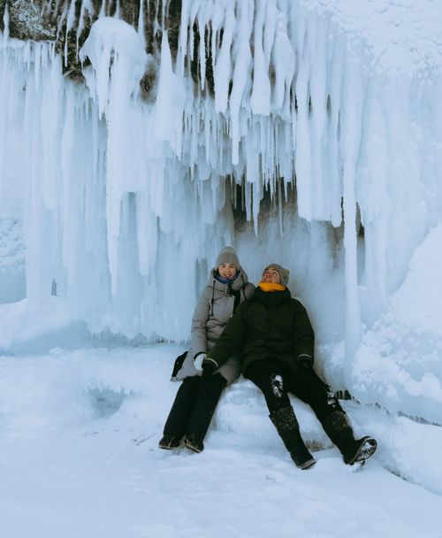 Free Couple sitting under icicles in winter, wearing warm clothing and jackets, enjoying the icy scenery. Stock Photo