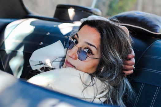 A fashionable woman with sunglasses relaxes inside a vintage car, exuding retro charm and style.
