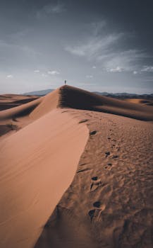 A lone person walks on sand dunes under a dramatic sky, showcasing vast desert landscape.