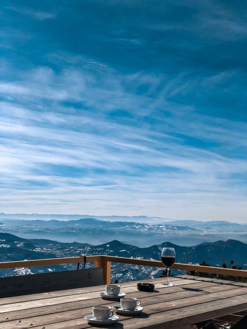 Free A peaceful mountain view with coffee cups and a wine glass on a wooden table under blue skies. Stock Photo