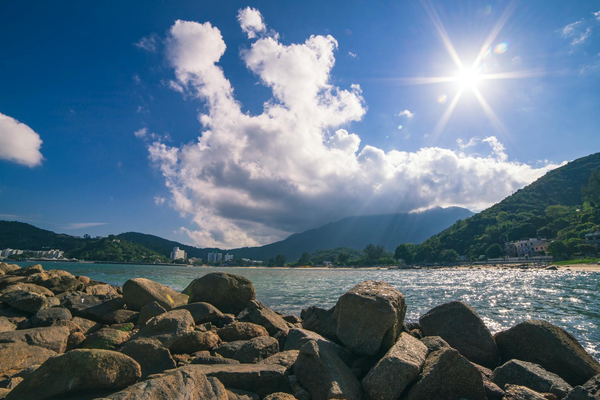 Dramatic sunny coastal view with rocky shoreline under bright blue sky and fluffy clouds.