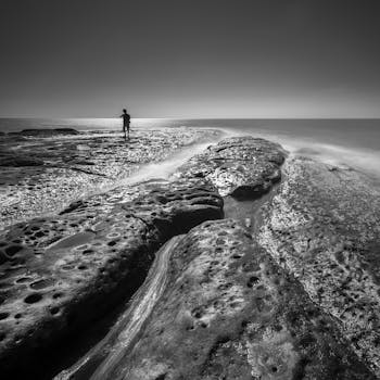 A black and white image showing a man silhouetted against an expansive rocky ocean seascape.