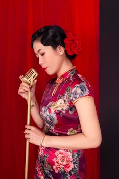 Asian woman elegantly singing in floral qipao, holding vintage microphone on red backdrop.