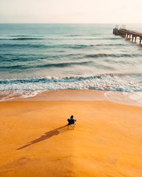 Man sitting alone on a sandy beach near waves, enjoying a tranquil sunset by the pier.