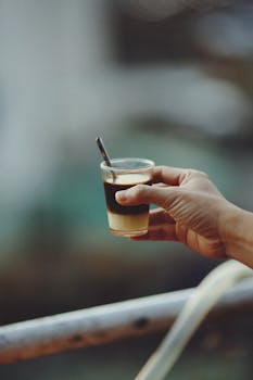 A close-up of a hand holding a layered coffee with a spoon outdoors, featuring soft focus.