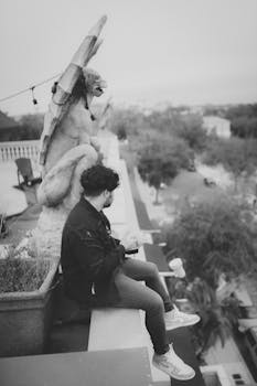 A man in a jacket sits by a gargoyle statue on a rooftop, captured in monochrome.