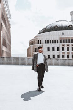 A fashionable man with sunglasses walks on a rooftop in an urban setting, exuding modern style.