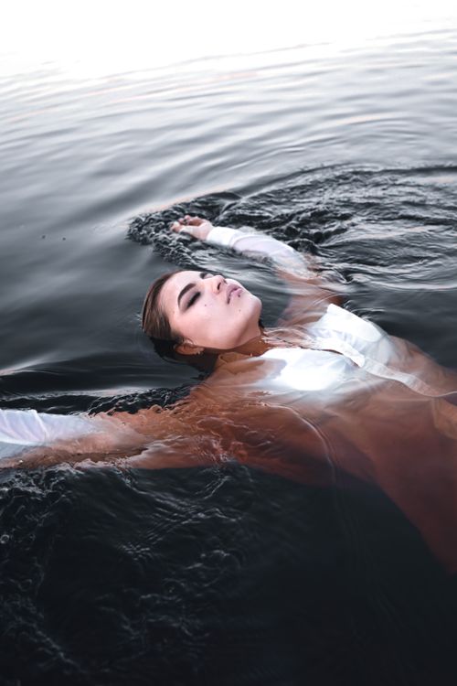 Free A woman in a white dress floats peacefully on dark water, eyes closed. Stock Photo