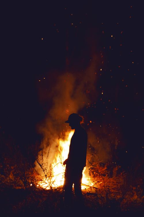 Free A person stands silhouetted against a blazing campfire, sparking a dramatic night scene. Stock Photo