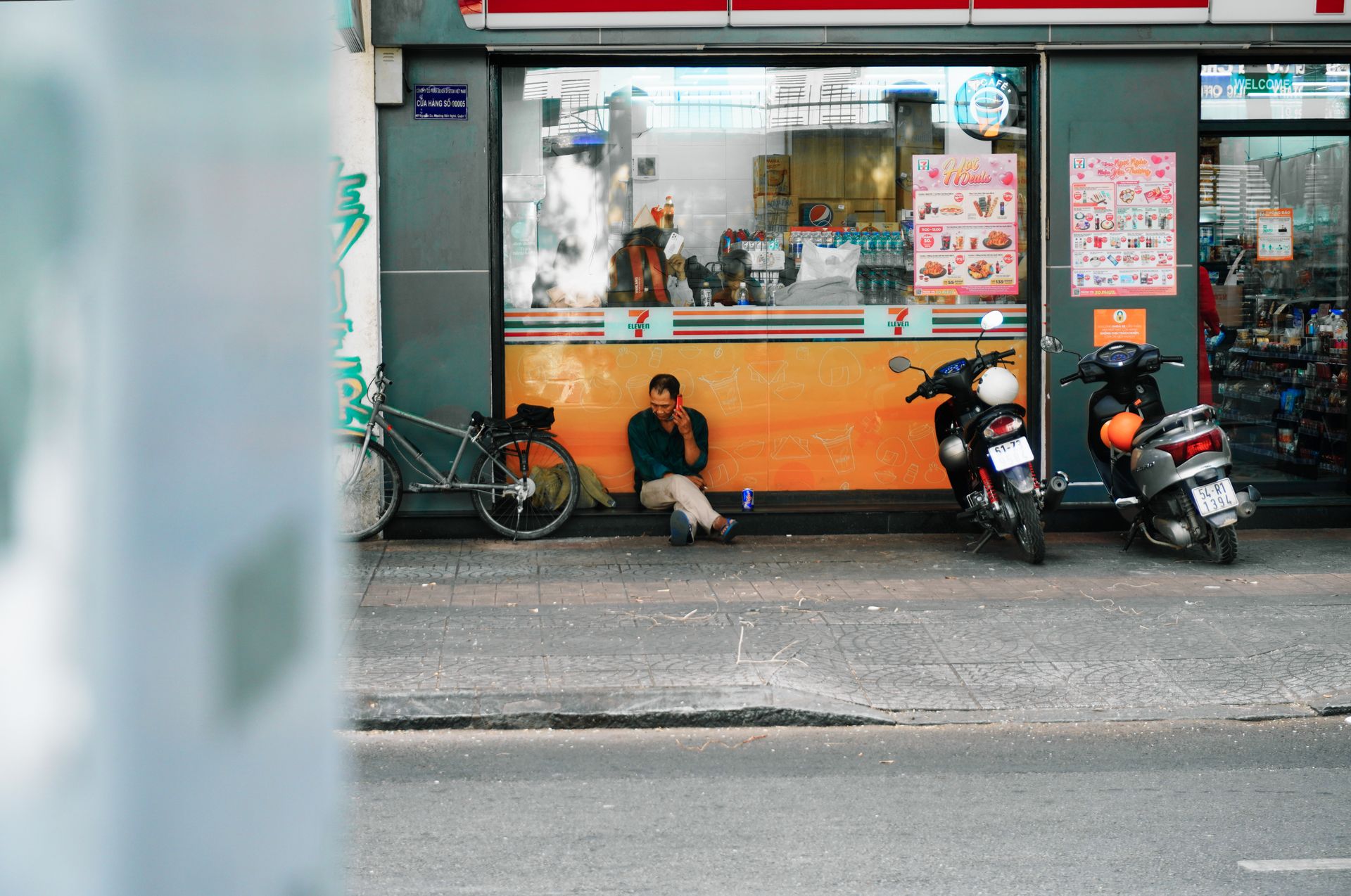 A person sitting in front of an urban convenience store with motorbikes parked nearby.