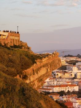 Dramatic sunset view of a town nestled against a rugged cliff with historic buildings.
