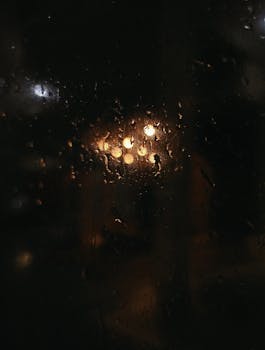 Raindrop-covered window capturing a warm glow and bokeh effect during nighttime.