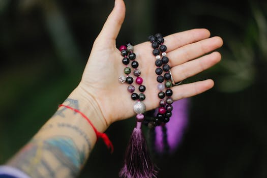 A tattooed hand holds vibrant Buddhist prayer beads against a lush green background.