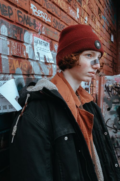 Free Young redheaded teenager in beanie and jacket, posing by graffiti wall with band aid on nose. Stock Photo