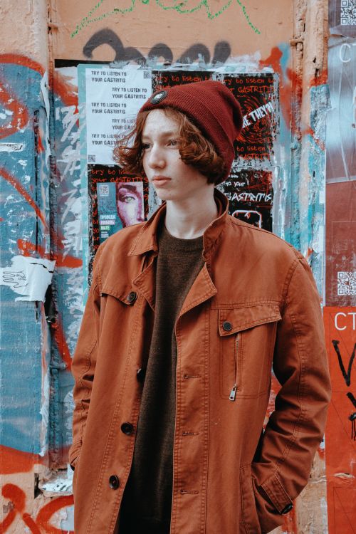 Free Stylish redhead teenager in urban setting wearing a jacket and beanie in front of graffiti-covered wall. Stock Photo