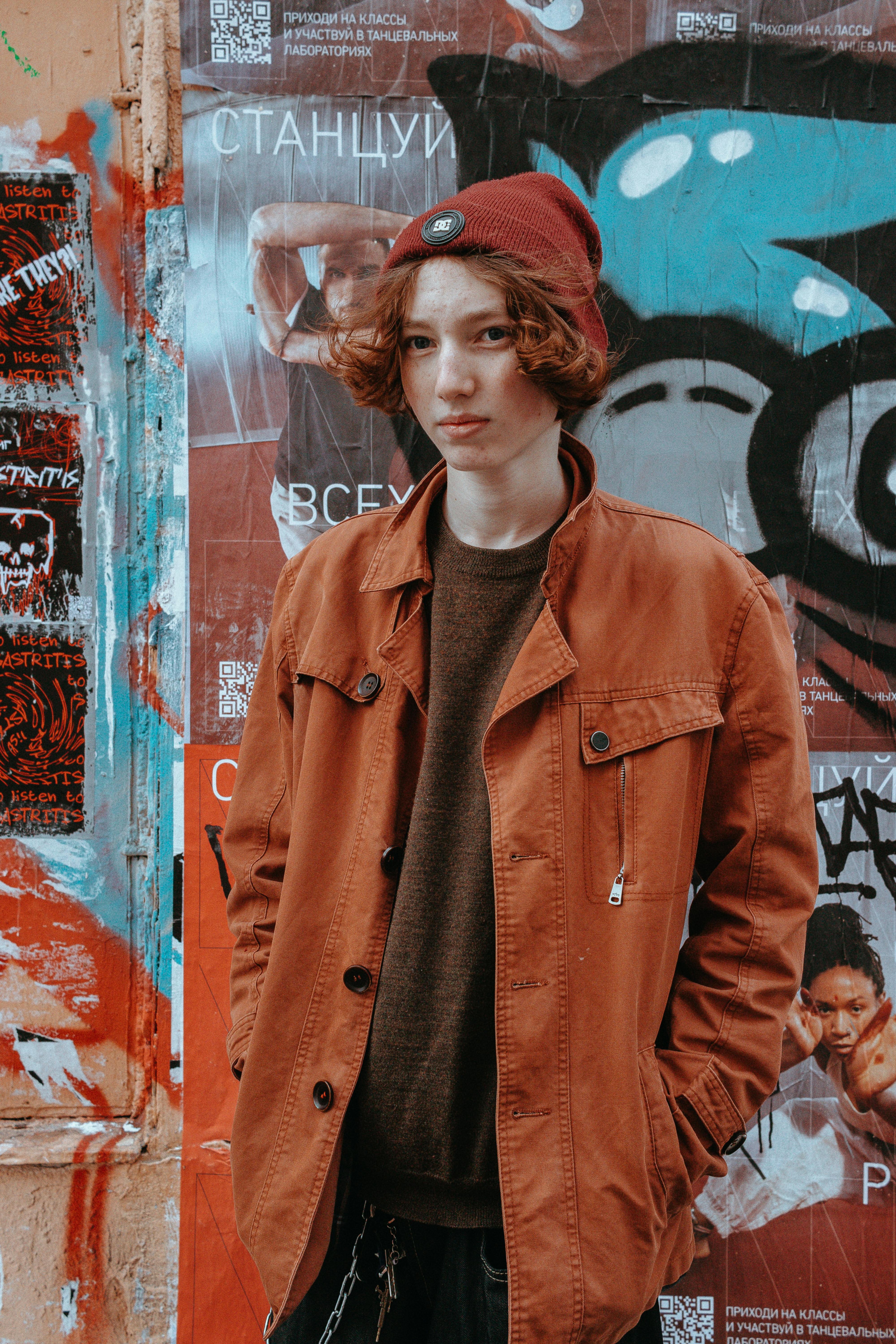 A young man in a brown coat and beanie stands against a graffiti-covered wall.