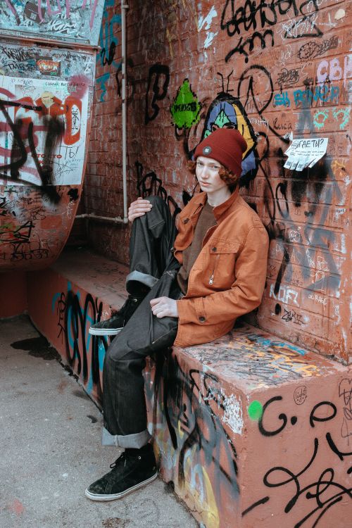 Free Teenager in fashionable attire sitting by a graffiti-covered wall, exuding modern urban style. Stock Photo