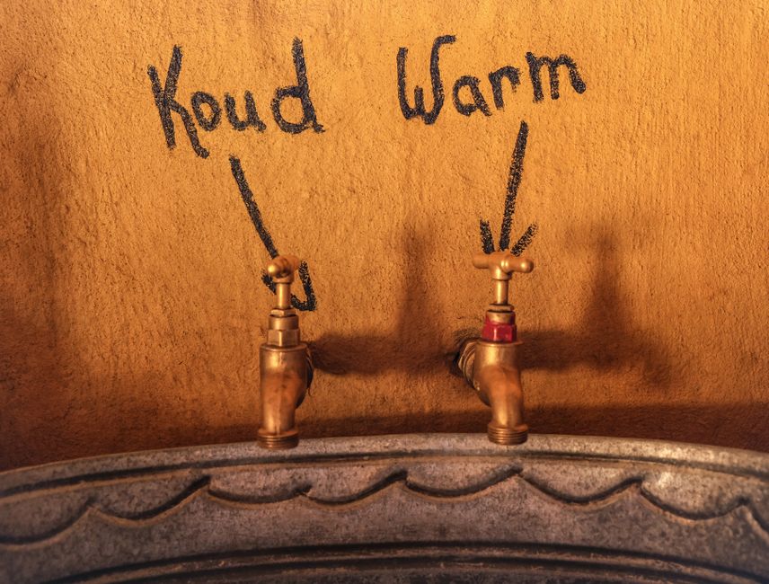 Photo by Magda Ehlers Close-up of vintage metal faucets labeled cold 'Koud' and warm 'Warm' on a textured gold wall.