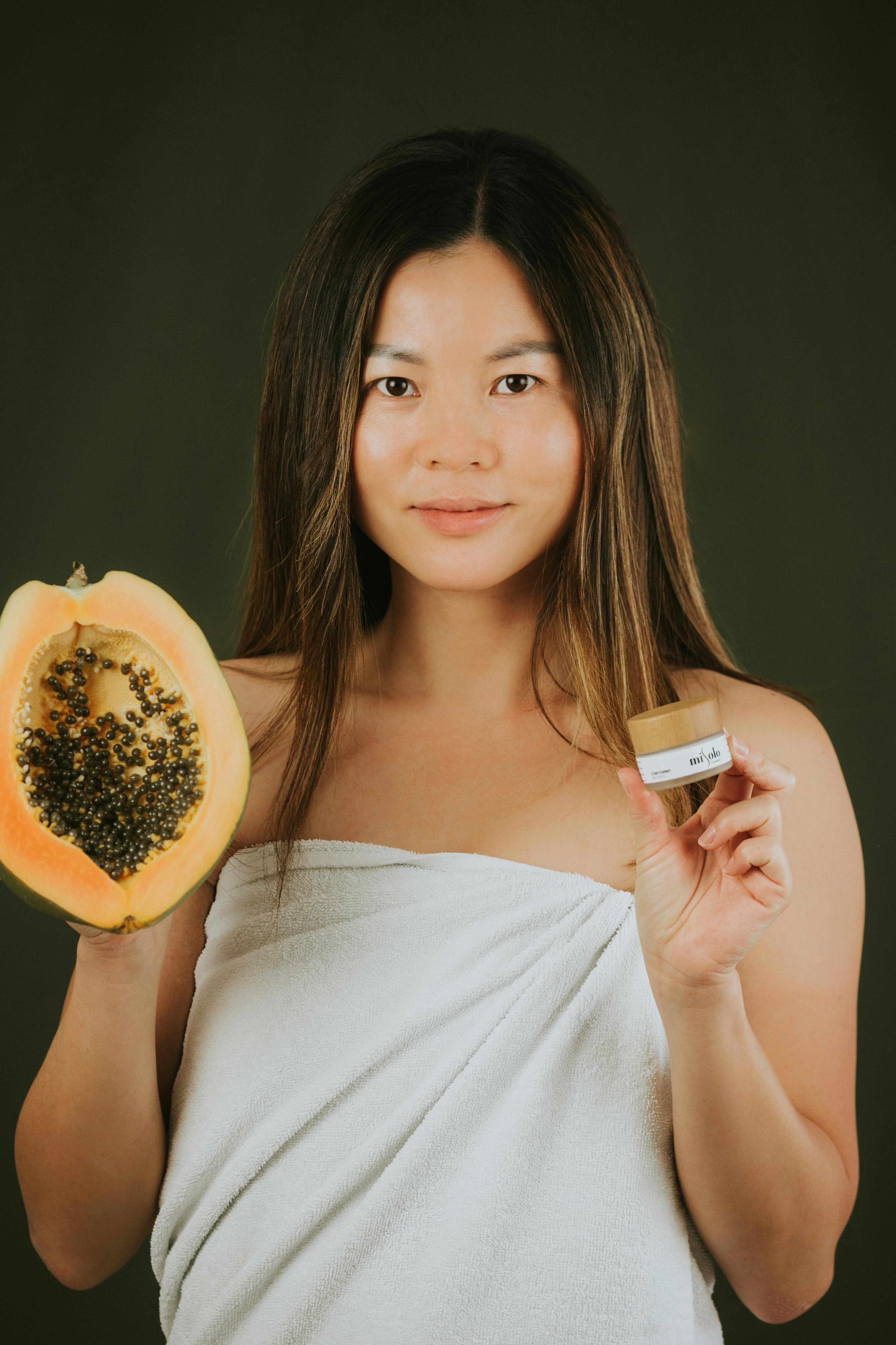Portrait of an Asian woman holding papaya and beauty product, promoting natural skincare.