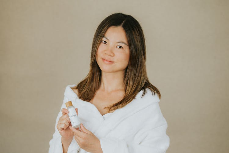 A Portrait Of A Woman In A White Robe Holding A Dropper Bottle