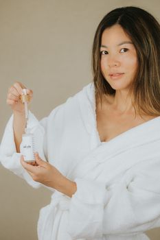 Asian woman in robe holding a skincare dropper bottle for beauty routine.