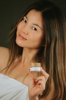 A beautiful Asian woman in a studio setting holding a skincare product while wrapped in a towel.