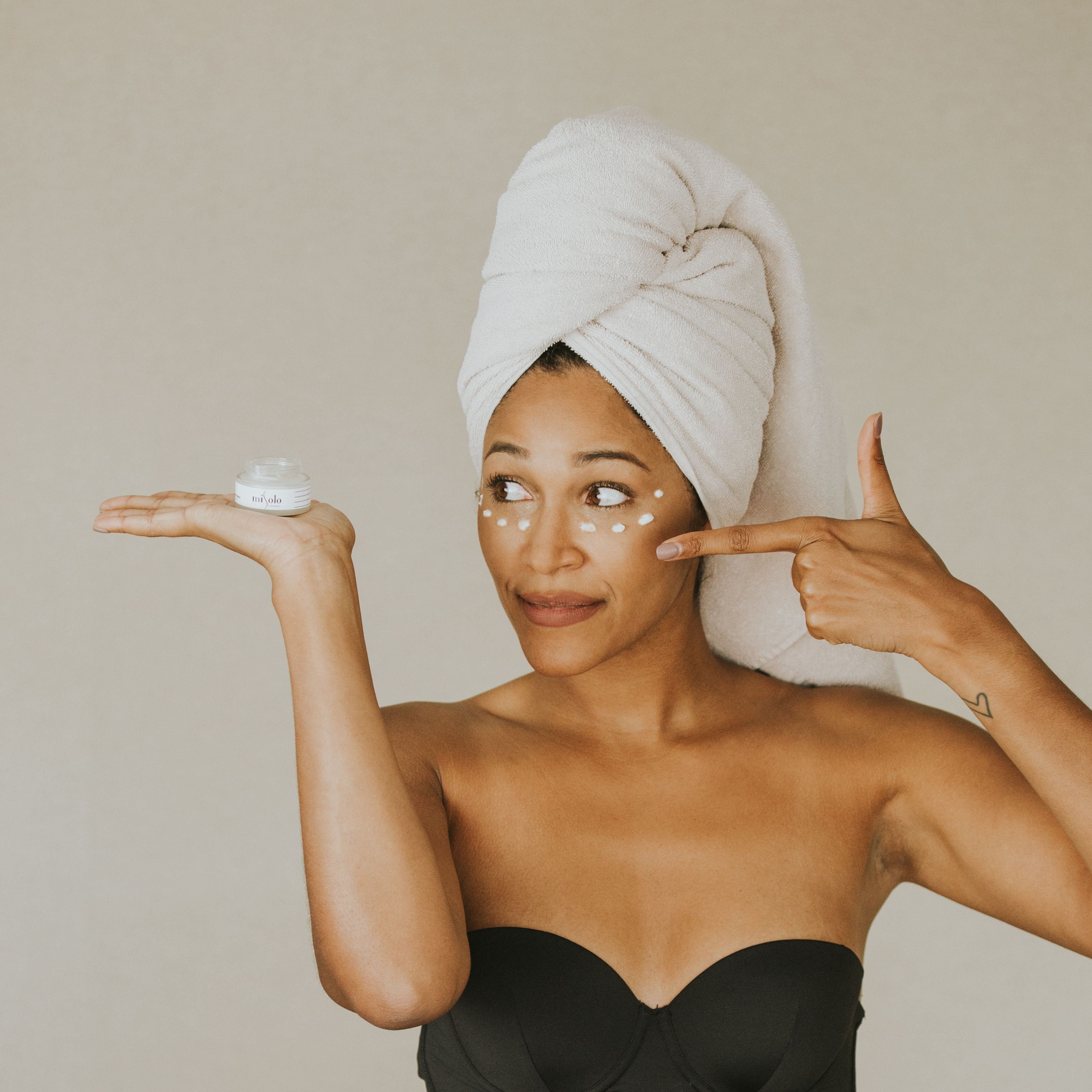 A woman with her hair wrapped in a towel. She has a skincare product in one hand and is pointing at it. 