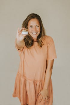 A cheerful woman in a peach dress presenting a face cream product indoors.