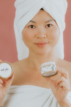 Asian woman with towel holding skincare products, promoting beauty routine.