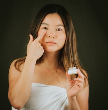 Asian woman applies skincare cream on face, focusing on beauty and self-care indoors.