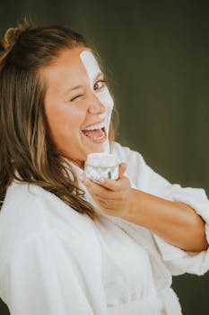Happy woman applying a skincare face mask while winking in a cozy setting.