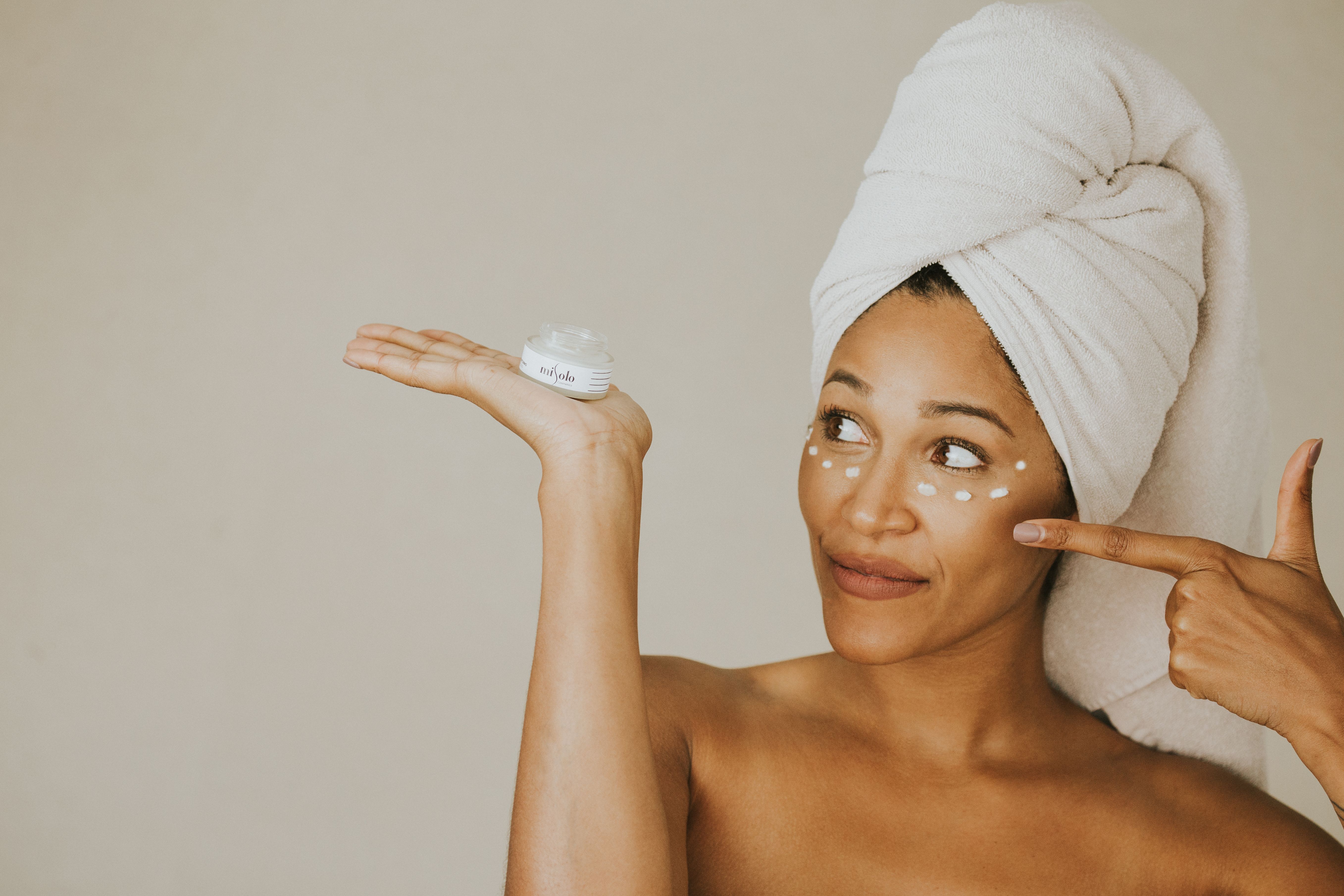 A woman applying face cream with a wrapped towel on her head, smiling.