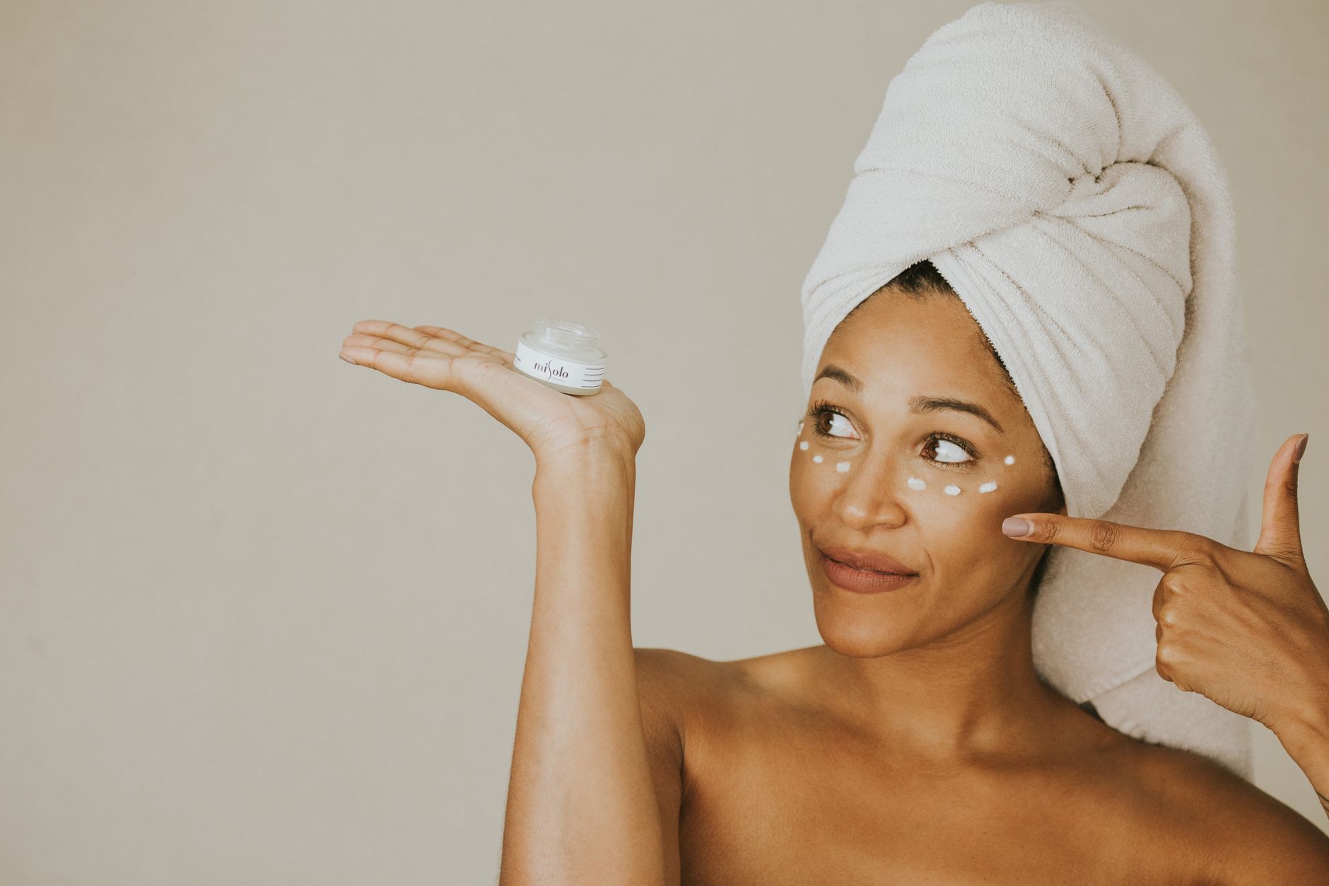 A woman applying face cream with a wrapped towel on her head, smiling.