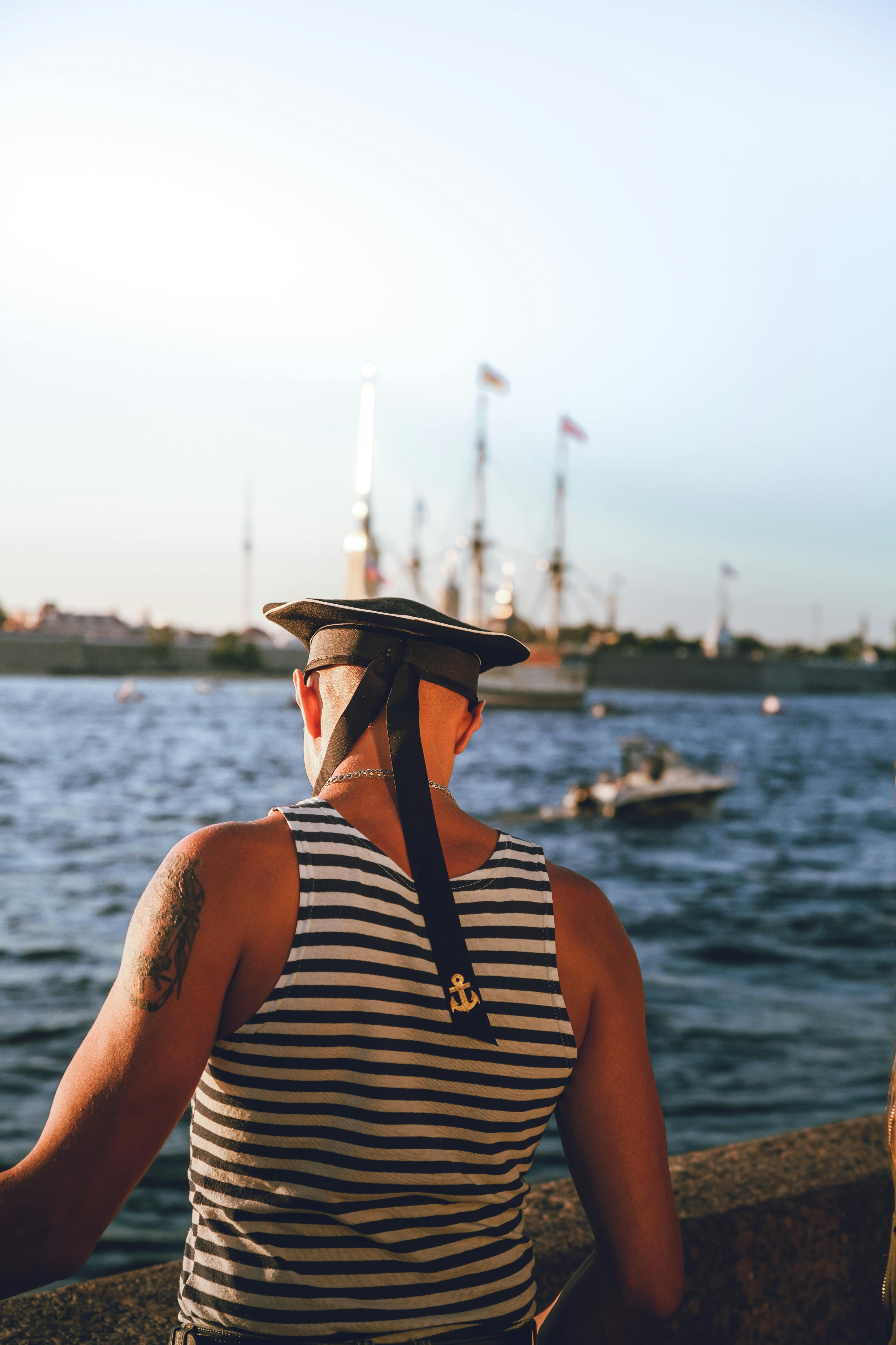 Tattooed man in sailor cap and striped tank top looking at boats on the water. Nautical and summer vibes.