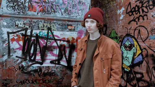 Free A young adult in a beanie and jacket stands against a vibrant graffiti wall. Stock Photo
