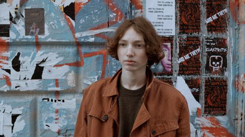 Free A young person wearing a brown coat stands near a colorful vandalized wall. Stock Photo
